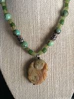 Close-up of carved soapstone pendant and green bead necklace on fabric background.