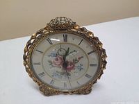 Front angle of vintage wind-up clock showing ornate gold-tone frame and floral petit point clock face with Roman numerals.
