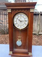 Full front view of wooden wall clock showing face with Roman numerals and visible pendulum inside glass case.