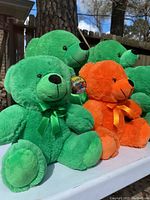 Many plush teddy bears in green and orange with ribbons and Colorama tags displayed outdoors on a white surface near trees.