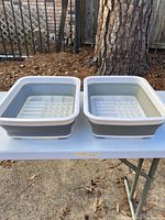 Two collapsible dish drying racks placed side by side on a white table outdoors with an outdoor fence and tree visible in the background.