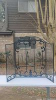 Full front view of black decorative metal fireplace guard on white table outdoors with background house and tree.