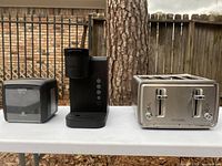 Photo showing Dixie Ultra napkin dispenser, black Keurig coffee maker, and stainless steel Cuisinart 4-slot toaster on white table outdoors against wooden fence.