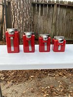 Four red storage containers with attached metal spoons arranged from largest to smallest on a white table outdoors, wood fence background.