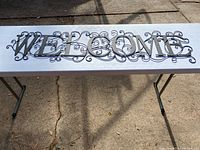 Full view of the large black metal 'WELCOME' sign placed on a white table outdoors on a paved surface, showing the entire sign and decorative scrollwork.