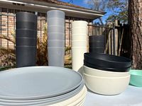 Stack of cups in black, gray, and cream, alongside stacked bowls and plates in neutral colors outdoors.