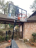 Clear acrylic backboard mounted on black aluminum frame with orange breakaway rim and white net, attached to black steel adjustable height pole and wheeled base outdoors near brick wall and wooden fence.