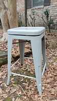 Full view of light gray metal swivel stool with square seat and straight legs placed outdoors on leaves.