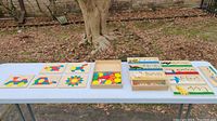 Table display of multiple wooden puzzle boards each with colorful geometric shapes arranged in patterns, plus a wooden box with assorted colored wooden shapes inside, set outdoors on white folding table.