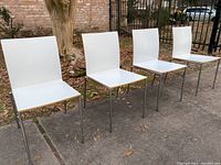 Four white dining chairs arranged in a row outside, showing the full chairs with metal legs and white seats and backs.