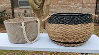 Two wicker baskets on a white table, one large round tan and black basket with handles, one light-colored basket with rope handles.