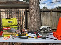 Full view of all tools on table including Ryobi circular saw in carrying case and box, hammers, drill, level, safety cones stacked, and other tools.
