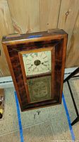 Front view of antique wooden framed wall clock with hand-painted tin dial and decorative panel. Keys visible on carpet below.