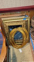Photo showing several ornate gold-painted wood frames stacked against a wall, including two oval frames at the front with detailed floral carvings, one with glass intact.