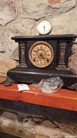 Front view of Seth Thomas windable mantle clock showing ornate bronze face and columns, cracked glass face not attached, winding key and plastic bag on shelf below clock, and round Westclox Baby Ben clock on top