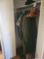 Closet view showing multiple green military coats and shirts hanging on wooden hangers, with a pair of worn boots below.