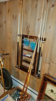 Six pool cues and two ping pong paddles displayed on a mounted wooden rack alongside billiard balls and other accessories.