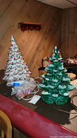 Two ceramic Christmas trees, green and white, with colorful bulbs and extra bulbs in plastic bags displayed on a table in a wood-paneled room.