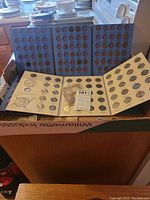 Photo showing two blue folders for penny collection and beige folders for state quarters with coins displayed, some currency notes in front