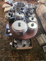 Lifetime brand stainless steel pots and pans with lids arranged on a table and a cardboard box beneath with more lids and cookware.