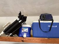 Photo showing two hard plastic coolers (one Coleman PolyLite 48) and one soft cooler bag along with folded black chair and table in garage shelf.