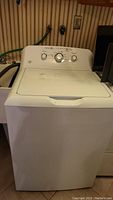 Front view of GE white top load washing machine with dial controls on the back panel and connected water hoses.