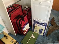 Three red soft-sided wheeled luggage pieces stacked with two wardrobe storage bags on floor.
