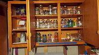 Wide view of wooden cabinet with multiple shelves containing assorted glass jars of various sizes and colors including clear and blue-green tinted glass.