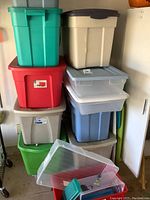 Stack of various colored plastic storage bins in garage including red, green, gray, beige, translucent blue bins with lids