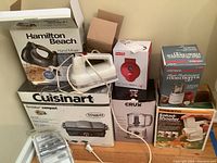 Photo showing Hamilton Beach hand mixer box, KitchenAid mixer with cord, Cuisinart Griddler Compact box, Presto Salad Shooter box, CRUX food chopper box, West Bend food chopper box, and other small appliance boxes stacked on floor.
