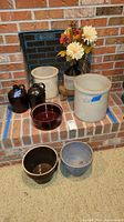 Photo showing multiple stoneware crocks and jugs, small wooden stool, bowls, and faux flowers arranged on a brick hearth.
