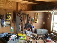 Wide view showing wooden pegboard with various hand tools hanging, a Craftsman circular saw in its carrying case, a metal tool box, hand tools including saw, brushes, and clamp. Some cleaning sprays on the workbench.