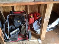 Photo showing red Toro push mower with fold-down handle, black engine, two red gas cans, and two cloth bags on wooden floor inside shed