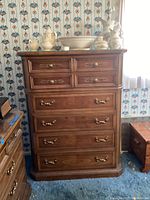 Full front view of wood chest of drawers showing all six drawers with brass handles and key escutcheons, placed against wallpapered wall and carpeted floor.