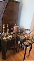 Overall grouping of brass candlestick holders, copper pot and kettle, brass urn, and enameled salt and pepper shakers on small wooden tables.