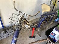 Front angle of the vintage Monark Super Deluxe bicycle showing the blue frame, cream seat, metal basket, and manual pump attached. The frame and components show significant rust, dirt, and wear.