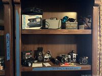 Photo showing vintage cameras and flashgun on wooden shelf: Polaroid cameras including Spectra 2 on box, gray Polaroid 210 with strap, vintage Kodak Brownie and Baby Brownie box cameras, Kuribayashi Petri camera and manuals.