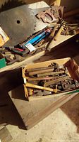 Photo shows wooden handles of several hacksaws, large circular saw blades and hand saws lying on a table.