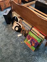 Photo showing vintage vinyl records leaning against a bed frame on carpet, with a black vinyl record carrying case beside them. Some visible albums include Village People and a record in a brown cover.