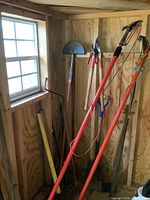 Photo of various garden tools hung on wall including pole pruner, shovel, pruning shears, axe with wooden handles and some stakes