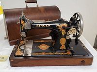 Front and side angled photo of the vintage Singer sewing machine on wooden base with ornate gold decals and curved wooden case in background.