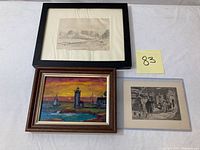 Front view of three artworks displayed on white surface, showing the lighthouse painting, landscape sketch, and street scene etching.
