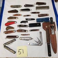 Top-down view of entire collection showing 27 vintage pocket knives, multi-tools, and a large fixed blade knife with sheath arranged on white cloth with blue edges.
