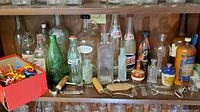 Full shelf view showing an assortment of vintage glass bottles with various labels and sizes, novelty swizzle sticks in red box, and several vintage bottle openers with wooden handles.