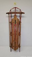 Full sled view showing wooden slats with red painted metal frame and rope handles, standing upright against a wall.