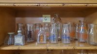 Shelf displaying 14 vintage glass bottles including various milk bottles and a toilet water bottle with label, some with original lids.