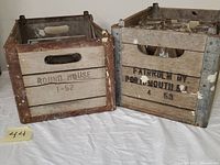 Two vintage wooden dairy crates side by side. Left crate labeled Round House 1-52, right crate labeled Fairholm Dairy Portsmouth R.I. 4 53, both weathered with metal corner reinforcements.