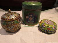 Three cloisonné jars shown together on fabric with different shapes and vibrant floral designs.