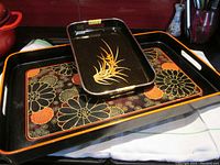 Two lacquered trays, one larger with floral orange and black pattern and one smaller black tray with floral motif, both with integrated handles