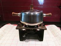 Full view of the stainless steel fondue pot with teak wood handles and knob atop the metal burner on a cloth surface.